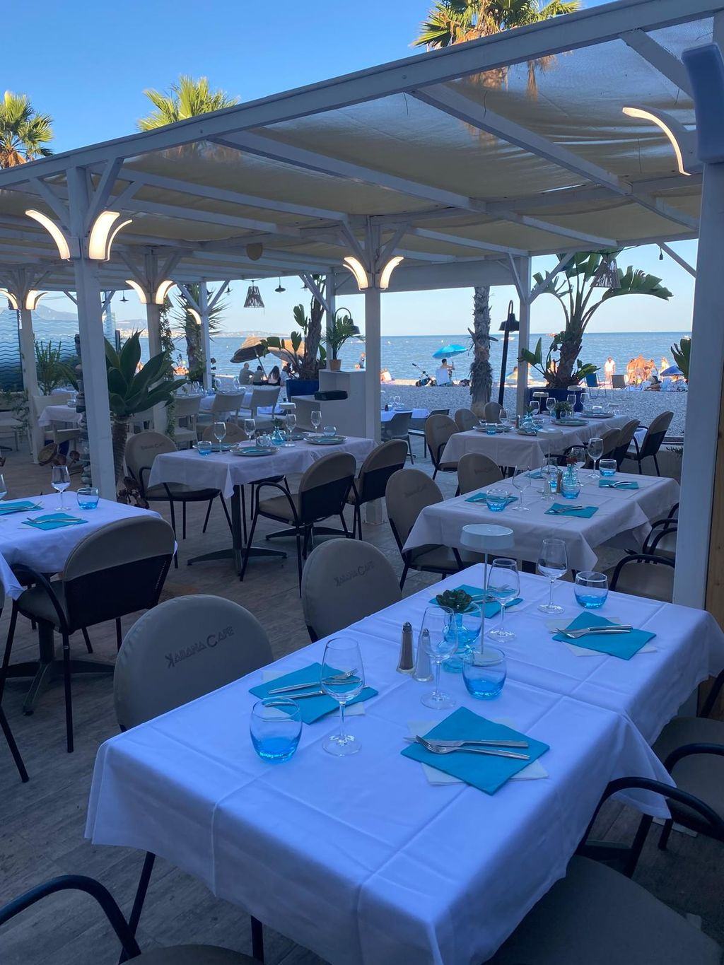 Tables dressées de nappes blanches et de serviettes turquoise sur la terrasse, en fin de journée, face à la mer